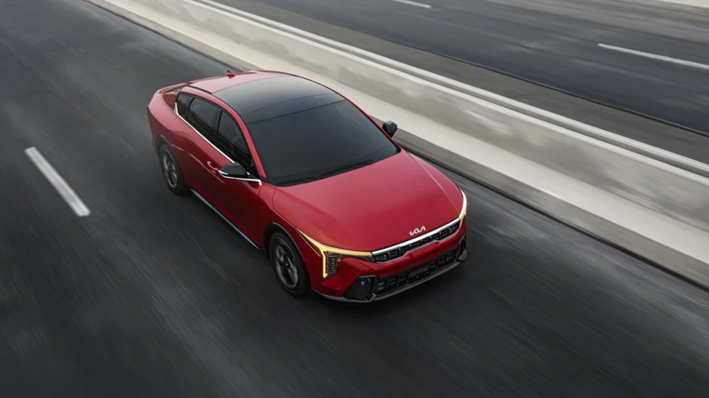 2025 Kia K4 in red, aerial front view, driving on highway with LED daytime running lights illuminated.