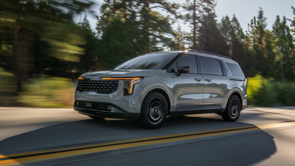 2025 Kia Carnival Hybrid in motion on a forest road showing sleek exterior styling and LED lighting.