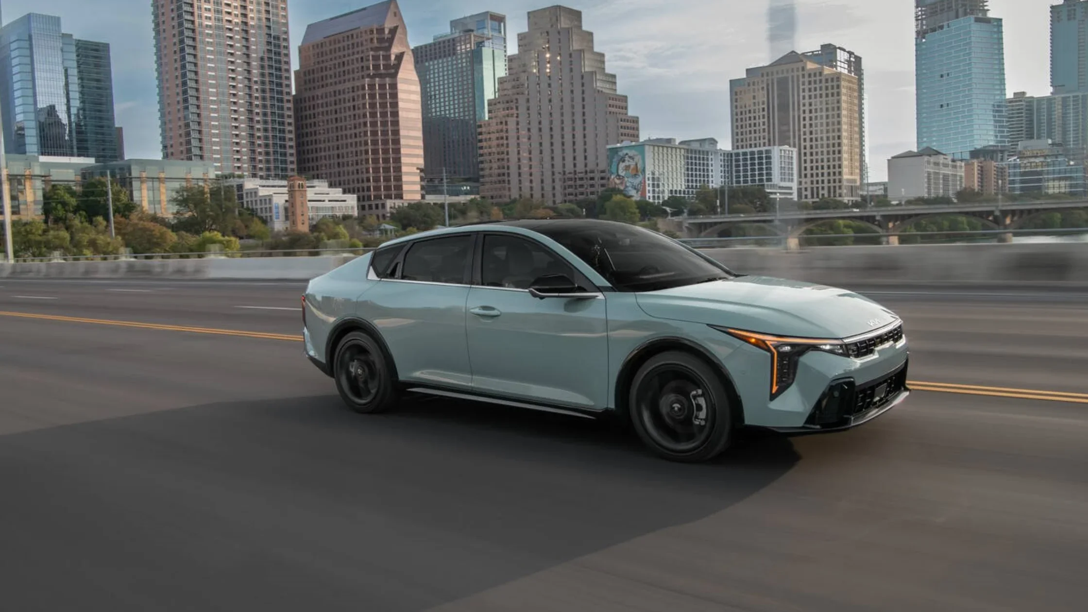 2025 Kia K4 sedan in light blue driving past modern city skyline with glass high-rises in the background.