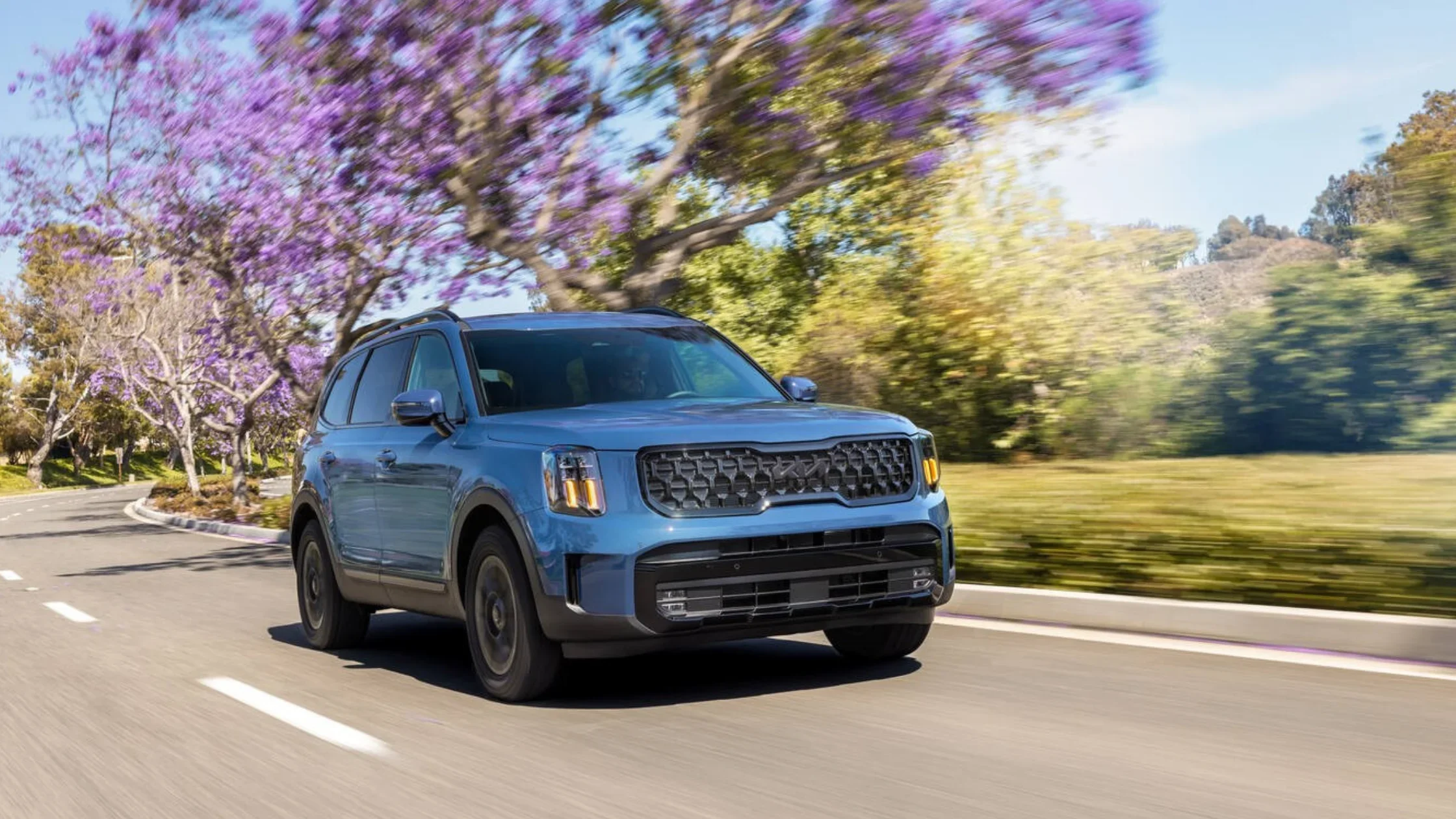 Blue 2025 Kia Telluride driving on a suburban road lined with trees and blooming purple jacarandas.