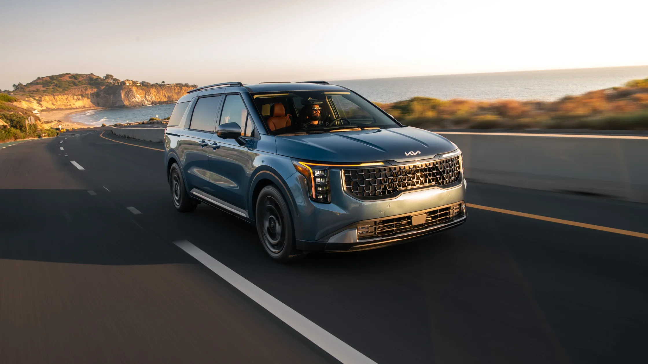 Blue 2025 Kia Carnival driving along a coastal highway at sunset with ocean cliffs behind.