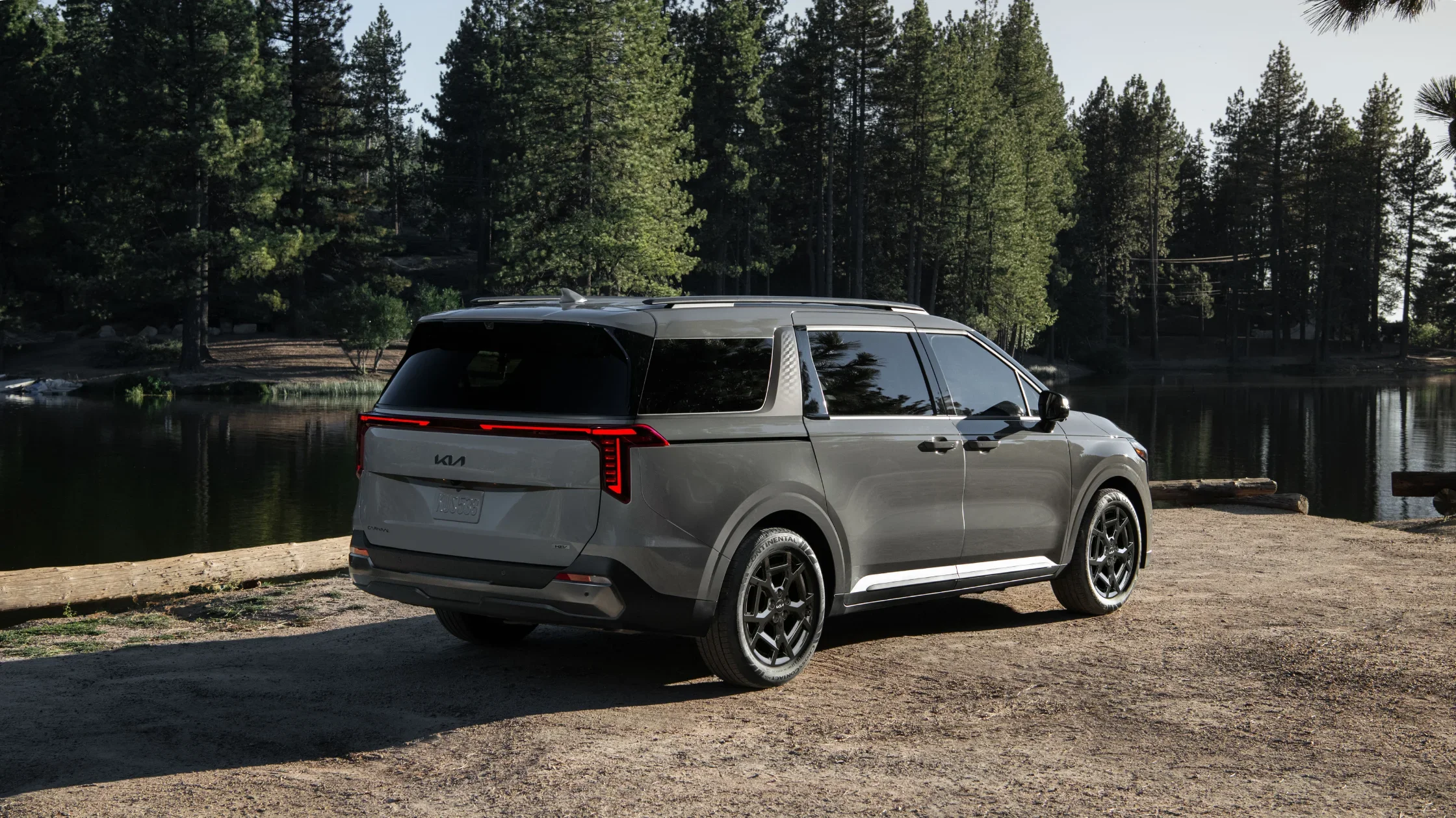 Gray 2025 Kia Carnival Hybrid parked lakeside with tall trees and calm water reflecting the minivan’s profile.