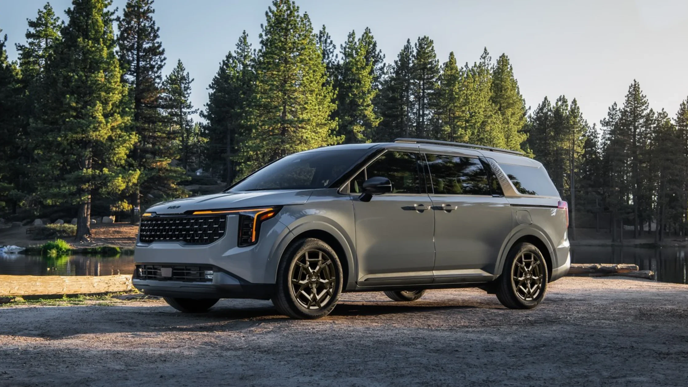 Side view of the 2026 Kia Carnival HEV parked near a scenic forest lake showcasing its sleek design.