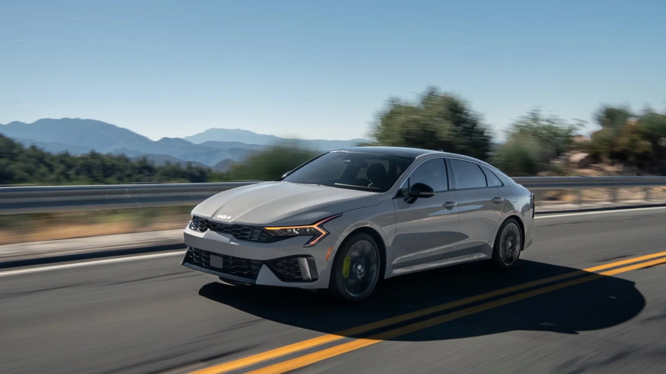 2026 Kia K5 sedan driving on a mountain highway showcasing aerodynamic styling and LED headlights.