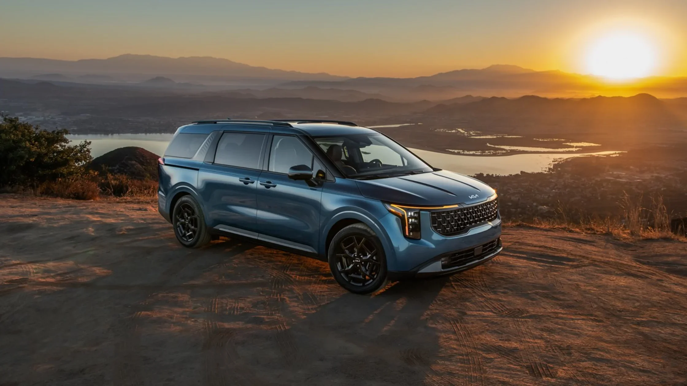 2026 Kia Carnival in blue parked at sunset on hillside overlooking valley and city lights.